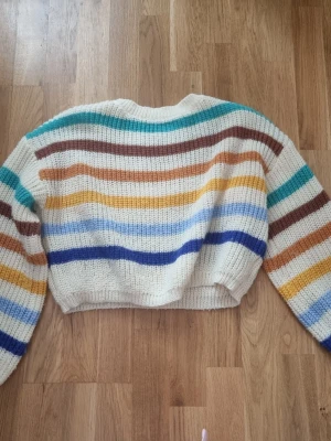 Randig stickad tröja  - Mysig stickad tröja i storlek L med breda ränder i vitt, blått, brunt, gult, orange och turkost. Tröjan har en croppad passform och långa ärmar. Perfekt för dig som gillar färg och vill ha en chill vibe i garderoben. Den är väldigt liten i storlek.