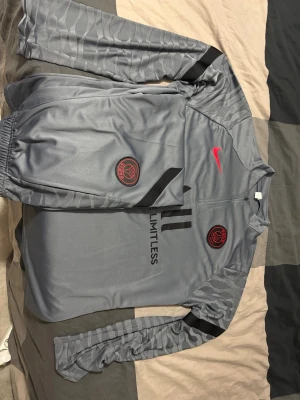 Grå PSG x Nike träningsset - Säljer ett grått PSG x Nike träningsset med långärmad tröja och matchande byxor. Setet har röda Nike-loggor, svarta detaljer och PSG-emblem på både tröja och byxa. Materialet är syntet och tröjan har half zip framtill. Perfekt för fotbollsträning eller chill.