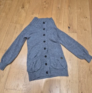 Grå stickad kofta från Burberry st S - Mysig grå stickad kofta från Thomas Burberry med hög krage, svarta knappar och två stora fickor framtill. Riktigt varm kofta av alpacka och merinoull. Klassisk logga i metall på ena fickan och ribbade muddar vid ärmslut och nederkant. Perfekt för kyliga dagar. I jättefint skick, knappt använd!
