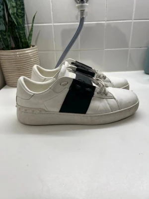 Valentino Garavani vita sneakers - Säljer ett par snygga vita Valentino Garavani sneakers med bred svart läderdetalj över snörningen och coola nitar längs hälen. Skorna har rund tå, platt sula och klassisk snörning. Märkeslogga i guld på plösen. Perfekt för dig som gillar exklusiva sneakers med unik design.