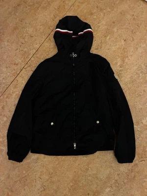 Svart vindjacka från Moncler - Säljer en svart vindjacka från Moncler med huva och dragkedja framtill. Jackan har vita och röda detaljer på huvan samt två fickor med tryckknappar. Perfekt för dig som vill ha en stilren och sportig look.