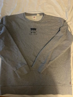 Grå sweatshirt från Levi's, storlek L - Klassisk grå sweatshirt från Levi's med tryck på bröstet där det står 'Levi's SF/CA EST. 1873'. Tröjan har rund halsringning, långa ärmar och ribbade muddar. Tillverkad i mjuk bomullsmix och har den ikoniska röda Levi's-taggen i sidan.
