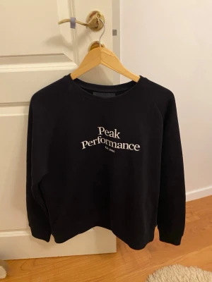 Svart Peak Performance sweatshirt - Svart sweatshirt från Peak Performance med vit logga framtill. Klassisk rund halsringning och långa ärmar. Perfekt basic-tröja i mjuk bomullsblandning som funkar året runt. Enkel och stilren design.