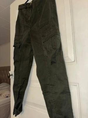 Olivgröna cargopants med knappar H&M - Säljer ett par olivgröna cargopants med stora sidofickor och snygga knappdetaljer vid bensluten från H&M. Byxorna har rak passform och är gjorda i bomull, perfekta för en chill och avslappnad stil.