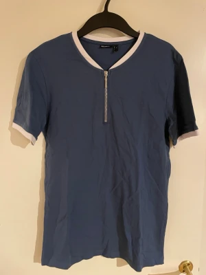 Blå t-shirt med dragkedja från ASOS - Blå t-shirt från ASOS DESIGN med vita detaljer vid krage och ärmslut. Kortärmad modell med en snygg dragkedja framtill. Tillverkad i mjuk bomull som känns skön mot huden. Perfekt för dig som gillar en clean och sportig look.