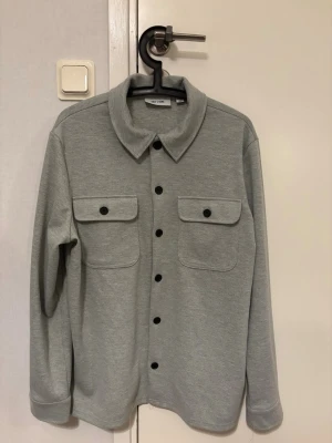 Grå overshirt från Only & Sons - Snygg grå overshirt från Only & Sons med klassisk krage, svarta knappar och två stora bröstfickor. Tillverkad i mjukt material som känns skönt mot huden. Perfekt lager-på-lager-plagg med avslappnad passform och lång ärm.