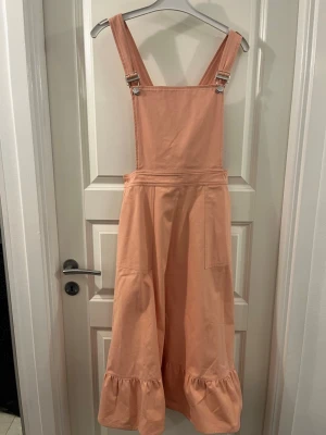 Ljusrosa långklänning från Wacay - Supersöt ljusrosa långklänning från Wacay med breda axelband och justerbara spännen. Klänningen har volangkant nertill och stora fickor framtill. Tillverkad i mjuk bomull, perfekt för sommarens alla äventyr.