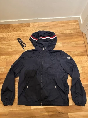 Marinblå vindjacka från Moncler - Snygg marinblå vindjacka från Moncler med huva och vita/röda ränder. Jackan har dragkedja framtill, två fickor med tryckknappar och ett broderat Moncler-märke på ärmen. Tillverkad i polyester och har en klassisk, sportig look. NFC tag funkar, snöre följer med.