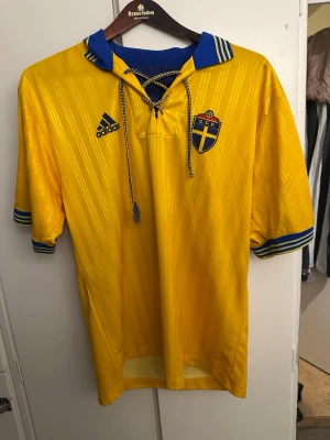 Gul vintage Sverige fotbollströja Adidas - Retro Sverige landslagströja från Adidas i gul och blå med klassiska ränder på ärmarna, snörning i halsen och S.F.F.-emblem på bröstet. Tillverkad i glansigt syntetmaterial och har randigt mönster i tyget. Perfekt för dig som älskar fotboll och old school-stil.