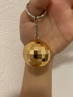 Guldig discokula nyckelring - Säljer en nyckelring med en liten discokula i guldigt plastmaterial. Kulan har spegelmönster som reflekterar ljus och ger en cool effekt. Perfekt accessoar till väskan eller nyckelknippan för dig som gillar lite extra bling.