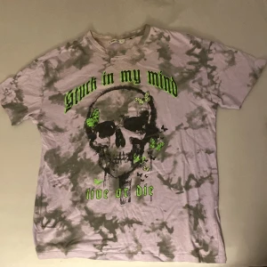 Tie dye t-shirt med dödskalle från FB Sister - Cool t-shirt från FB Sister i tie dye-mönster med grått och lila. Framsidan har tryck med dödskalle, gröna fjärilar och texten 'Stuck in my mind, live or die' i neon. Klassisk passform och korta ärmar. Perfekt för dig som gillar edgy streetstyle. Lite oversized S, känns som medium
