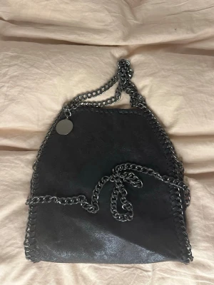 Svart axelväska med kedjedetaljer - Snygg svart axelväska i mockaimitation med kraftiga silverfärgade kedjor som axelband och kantdetalj. Väskan har magnetlås och insidan är fodrad med leopardmönstrat tyg samt har en innerficka med dragkedja. Perfekt för dig som gillar edgy detaljer och djurmönster.