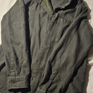 Mörkgrön overshirt i ullmix - Säljer en mörkgrön overshirt med klassisk krage och bröstficka. Skjortan har knäppning framtill och långa ärmar med manschett. Materialet är en mjuk ullmix som ger en skön och avslappnad känsla. Perfekt att bära lager på lager under kyligare dagar.