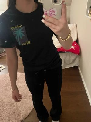 Svart t-shirt med palmtryck - Svart t-shirt från Hummel med tryck på bröstet som visar en blå palm och texten 'Sakana River' i gult. Klassisk passform och rund hals. Perfekt till chill dagar och enkel att matcha med jeans eller shorts.