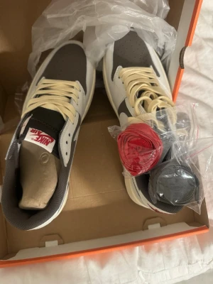 Nike Air Jordan 1 Low Travis Scott - Nike Air Jordan 1 Low Travis Scott i vitt, brunt och beige med ikonisk omvänd swoosh och röda broderade detaljer på hälen. Skorna har snörning och kommer med extra skosnören i rött och svart. Tillverkade i skinn och mocka för en clean och exklusiv vibe. Aldrig använda. Jag fick de i present men de är för stora för mig 
