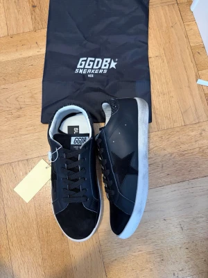 Svarta GGDB sneakers med glitterdetalj - Svarta GGDB sneakers med vit sula, svart stjärna på sidan och glittrig häl. Ovandel i skinn och mocka, snörning framtill och klassisk låg profil. Perfekta för dig som vill ha en clean men ändå unik look.