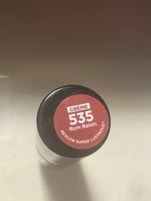 Revlon Super Lustrous Läppstift 535 Rum Raisin - Jättepopulär 90s färg, Revlon Super Lustrous Lipstick i färgen 535 Rum Raisin är ett krämigt läppstift i en djup bärig brunröd ton. Hylsan är svart med en elegant gulddetalj och logga. Formulan är återfuktande och ger en snygg, klassisk finish. Perfekt för dig som gillar naturliga men ändå markerade läppar.