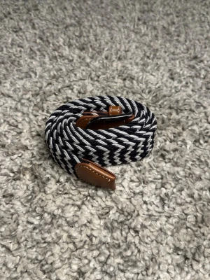 Flätat Bälte  - Svartvitt flätat bälte med bruna skinn-detaljer och metallspänne. Bältet har ett snyggt zigzagmönster och är elastiskt för extra komfort. 105cm långt