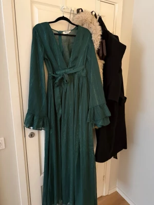 Grön långklänning från Showpo - Säljer en smaragdgrön långklänning från Showpo med subtilt glittrigt tyg, v-ringning och volangdetaljer vid ärmsluten. Klänningen har lång ärm, knytband i midjan och dragkedja bak. Perfekt för dig som vill sticka ut med en elegant look.               