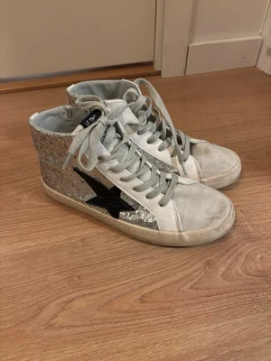Glittriga high top sneakers - Säljer ett par vita high top sneakers från Le Temps des Cerises med silverglittriga sidor och en svart stjärna på utsidan. Dom är sparsamt använda och har lite smuts längs fram som går att tvätta bort!