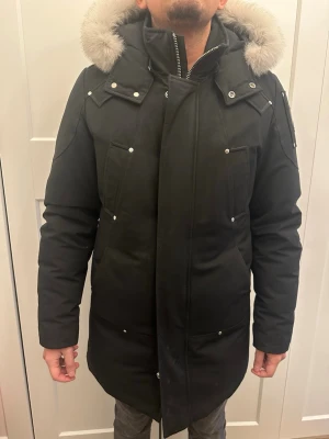 Svart parkas från Moose Knuckles - Säljer en svart parkas från Moose Knuckles med stor luva och fluffig pälsdetalj. Jackan har flera fickor med tryckknappar, dragkedja framtill och snygga metalldetaljer på ärmen. Perfekt för kalla vinterdagar och riktigt varm. Använd fåtal gånger