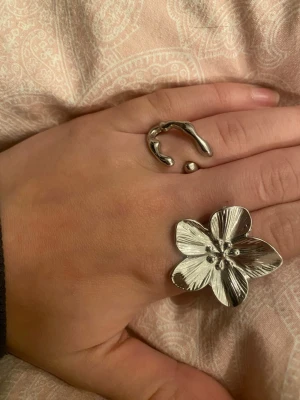 Snygga silvriga ringar med blomma - Två unika ringar i silverfärg, en med stor blomformad detalj och en med organisk, öppen design. Perfekta för dig som vill sticka ut och addera något extra till din stil. Båda ringarna är i metall och har en modern känsla.
