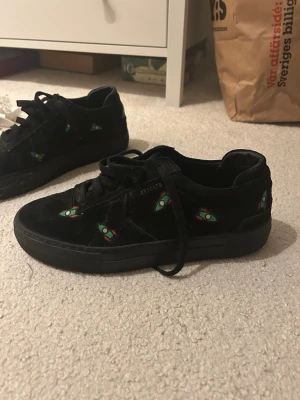 Svarta sneakers från Axel Arigato - Säljer ett par svarta limited edition sneakers från Axel Arigato i mocka med broderade små raketer i grönt och rött över hela skon. Skorna har snörning och en platt sula. Nypris är runt 2500-3000😻