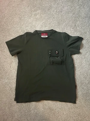 Mörkgrön t-shirt från Parajumpers - Säljer en mörkgrön t-shirt från Parajumpers i storlek S. T-shirten har en unik bröstficka med dragkedja och lock, samt Parajumpers-logga på fickan. Klassisk rund hals och korta ärmar. Perfekt för dig som gillar streetwear och funktionella detaljer.