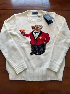 Vit stickad tröja Polo Ralph Lauren - Säljer en vit stickad tröja från Polo Ralph Lauren med ikonisk björn i röd kavaj broderad på framsidan. Tröjan har rund hals, ribbade muddar och är långärmad. Perfekt för dig som vill ha något klassiskt men ändå lekfullt. Ganska liten i storleken, passar någon runt 165
