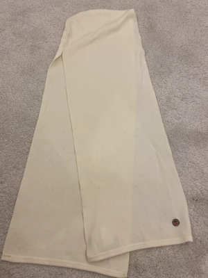 Beige väst i bomull med liten patch - Säljer en enkel och stilren beige halsduk i mjukt bomullstyg. Halsduken har rak passform och en liten rund patch längst ner som detalj (Busnel). Perfekt att styla över en t-shirt eller skjorta för en clean look.