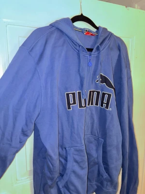 Blå puma hoodie med dragkedja - En blå hoodie från Puma med huva, dragkedja och snörning. En svart/vit puma logga över bröstet. 
