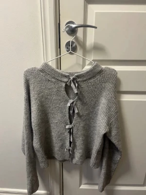 Grå stickad tröja med knyt från Gina Tricot - Mysig grå stickad tröja från Gina Tricot med långa ärmar och snygga knytband längs ryggen. Tröjan har en ribbad struktur och en relaxed passform som passar perfekt till höst och vinter. Enkel att styla med jeans eller kjol.