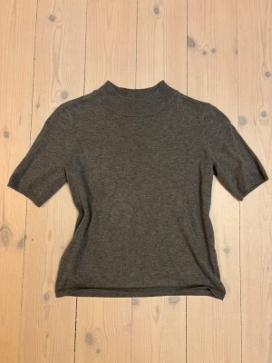 Grå stickad topp från Stockh LM - En stilren grå stickad topp från Stockh LM med korta ärmar och högre ribbad hals. Toppen har en rak passform och är tillverkad i mjukt material som känns skönt mot huden. Perfekt att matcha med både jeans och kjol.