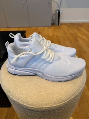Nike Presto helvita sneakers - Nike Presto sneakers i helvit färg med mesh-ovandel och syntetiska detaljer. Skorna har rund tå, platt sula och tydlig snörning. Perfekta för dig som gillar stilrena och sportiga vibes. Diskret Nike-logga på sidan och framtill.