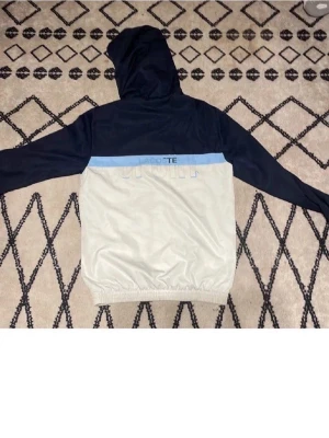 Lacoste vindjacka med huva - Snygg tracksuit från Lacoste i marinblått och vitt med ljusblå detalj över bröstet. Jackan har huva, hel dragkedja framtill och elastisk nederkant. Perfekt för dig som gillar sportig stil och vill ha något lätt och bekvämt. Byxorna är lika bekväma att röra sig i många olika sätt från att löpa till att spela fotboll med dom funkar 