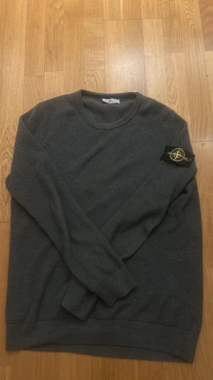 Mörkgrå stickad tröja Stone Island - Säljer en mörkgrå stickad tröja från Stone Island med klassisk patch på ärmen. Tröjan har rund halsringning och långa ärmar. Perfekt för dig som gillar streetwear och vill ha något stilrent men ändå med edge.