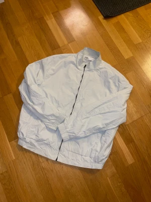 Vit vindjacka från Zara - Säljer en clean vit vindjacka från Zara i storlek L. Jackan har hel dragkedja framtill, hög krage och två stora fickor. Materialet är lätt och känns perfekt för blåsiga dagar. Resår vid ärmsluten ger en skön passform och enkel stil.