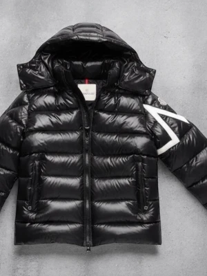 Moncler jacka - Säljer en svart pufferjacka från Moncler med huva och dragkedja. Jackan har ett stort vitt Moncler-märke på ena ärmen och två fickor med dragkedja framtill. Glansigt material och klassisk Moncler-stil, perfekt för kalla dagar.