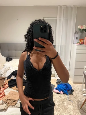 Svart spetsdetaljerad linnetopp - Säljer en svart linnetopp med smala axelband och snygga spetsdetaljer vid urringningen. Toppen har knyt i nacken och en v-ringad front som ger en feminin look. Perfekt att styla med jeans eller kjol för en trendig outfit.