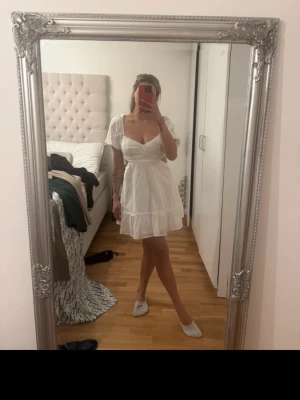 Vit kort klänning med volang - Superfin vit kort klänning med broderade detaljer och volang nertill. Klänningen har v-ringning, korta ärmar och markerad midja. Perfekt för sommarens alla tillfällen och ger en fräsch och romantisk look.