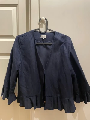 Mörkblå kofta med volang från Isolde - Snygg mörkblå kofta från Isolde i storlek 36. Koftan har långärmade ärmar och volangdetaljer längs nederkanten och ärmsluten. Tillverkad i ett linneliknande material och har en öppen front utan knappar eller dragkedja. Perfekt för att addera lite extra stil till din outfit.