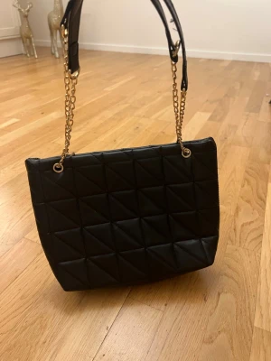 Svart quiltad axelväska med kedja - Snygg svart axelväska i quiltad design med guldfärgade kedjor och axelband. Väskan har en rymlig insida och stilren look som passar till många outfits.