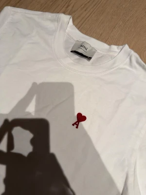 Vit t-shirt från Ami med rött hjärta - Vit t-shirt från Ami med klassisk passform och korta ärmar. På bröstet finns ett broderat rött hjärta med bokstaven A. Tillverkad i mjuk bomull och har en enkel, stilren look som är lätt att matcha.