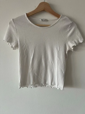 Vit ribbad croppad topp från ONLY - Säljer en vit ribbad topp från ONLY med korta ärmar och croppad passform. Toppen har en rund halsringning och vågig kant längst ner och vid ärmsluten. Perfekt till jeans eller kjol för en enkel och fräsch look.