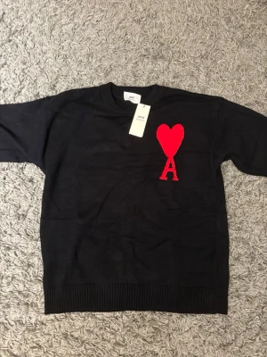 Svart stickad tröja från Ami Paris - Svart stickad tröja från Ami Paris med röd hjärt- och A-detalj på bröstet. Klassisk rund halsringning och ribbade muddar. Tillverkad i Portugal av mjuk ull som känns riktigt skön mot huden. Perfekt för dig som vill ha en stilren men ändå unik look.