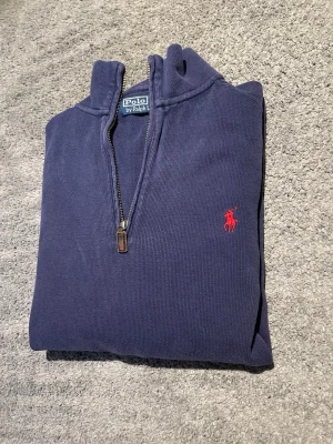 Mörkblå stickad tröja Polo Ralph Lauren - Mörkblå stickad tröja från Polo Ralph Lauren med klassisk röd logga broderad på bröstet. Tröjan har rund halsringning, långa ärmar och ribbade muddar. Perfekt för lager-på-lager och snygg till jeans eller chinos.