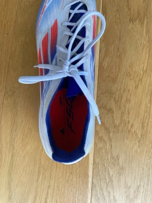 Adidas F50 blåvita fotbollsskor - Säljer ett par Adidas F50 fotbollsskor i vitt med blå och röda detaljer. Skorna har snörning, låg profil och mönstrad ovandel i syntetmaterial. Yttersulan har fasta dobbar för bra grepp på planen. Perfekta för dig som vill sticka ut på matchen!