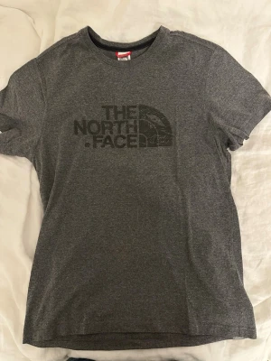 Grå t-shirt från The North Face - Säljer en grå t-shirt från The North Face med svart logotyp tryckt på bröstet. T-shirten har rund hals och korta ärmar, tillverkad i mjuk bomull som är skön mot huden. Perfekt för dig som gillar enkel och stilren streetwear eller som träningströja.