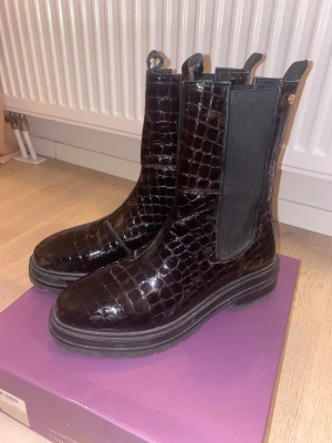 Bruna chelsea boots från Copenhagen Shoes - Säljer ett par snygga bruna chelsea boots från Copenhagen Shoes i storlek 41. Skorna har ett glansigt, krokodilpräglat skinn och elastiska paneler på sidorna för enkel på- och avtagning. Grov platt sula och rund tå ger en cool och modern look.