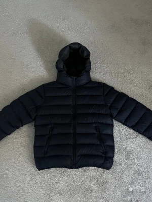 Mörkblå pufferjacka från Colmar - Säljer en mörkblå pufferjacka från Colmar med huva och dragkedja framtill. Jackan har ett stilrent, quiltat mönster och två fickor med dragkedja. Har en liten lagning i en ärmen. Pris kan diskuteras 